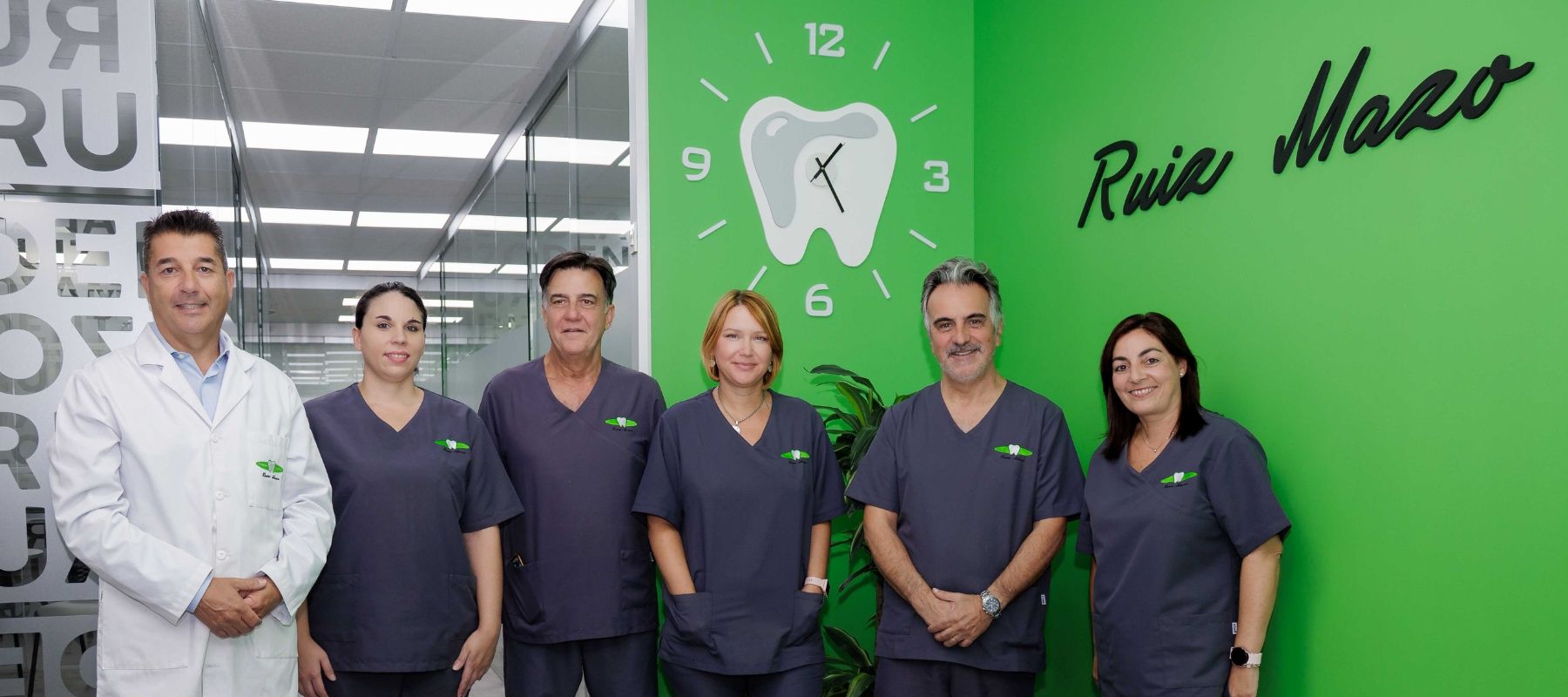 Seis profesionales de odontología posan en un pasillo de la clínica, delante de una pared verde con reloj en forma de diente.